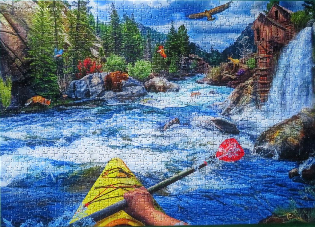 A first-person view from a kayak on a rushing river flowing past an old wooden mill and waterfall in a forest.