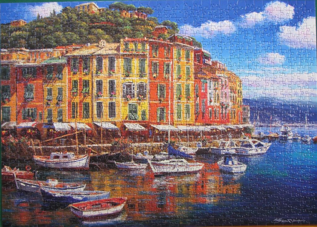 A coastal Mediterranean village with colourful buildings, boats in the harbor, and a lush green hillside.