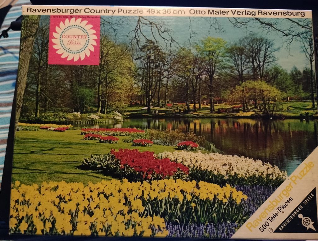 A lush spring garden with colorful flower beds beside a tranquil pond and tall trees in the background.