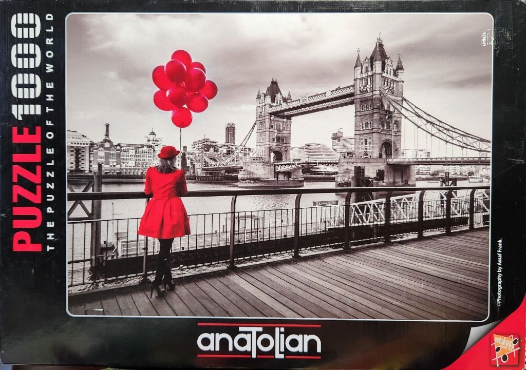 A woman in a red coat holding red balloons stands on a boardwalk looking toward Tower Bridge in black and white.