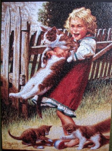 A young girl with blonde curly hair happily holding a large cat in front of a rustic wooden fence.