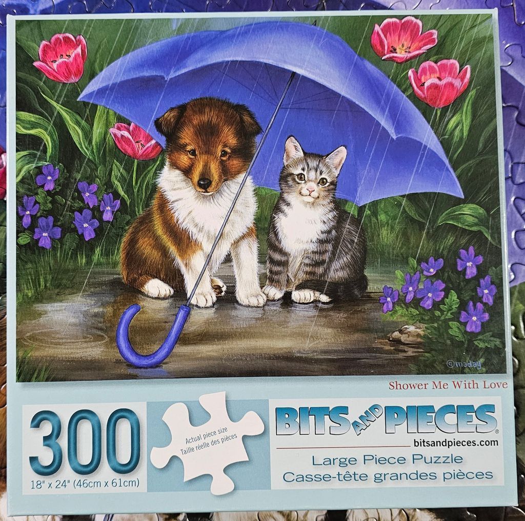 A puppy and a kitten sitting together under a blue umbrella in the rain surrounded by flowers.