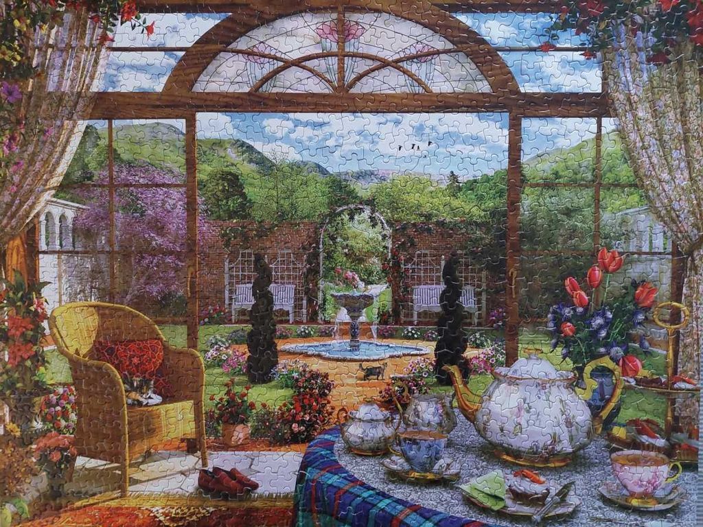 A sunroom overlooking a lush garden with a fountain, featuring a tea set on a table and a wicker chair.