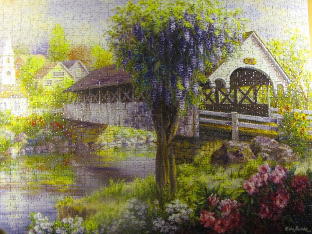 An idyllic covered wooden bridge over a stream surrounded by wisteria and blooming flowers.
