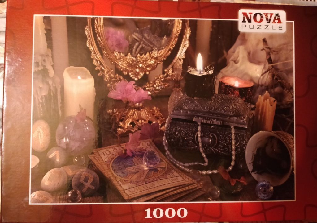 A still life featuring a gold mirror, a lit black candle on a decorative box, tarot cards, and runestones.