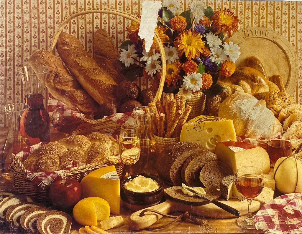 A bountiful still life featuring assorted breads, wheels of cheese, glasses of wine, and a bouquet of autumn flowers.