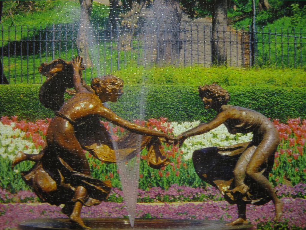 Two bronze statues of women dancing in front of a colorful flower bed and a fountain.