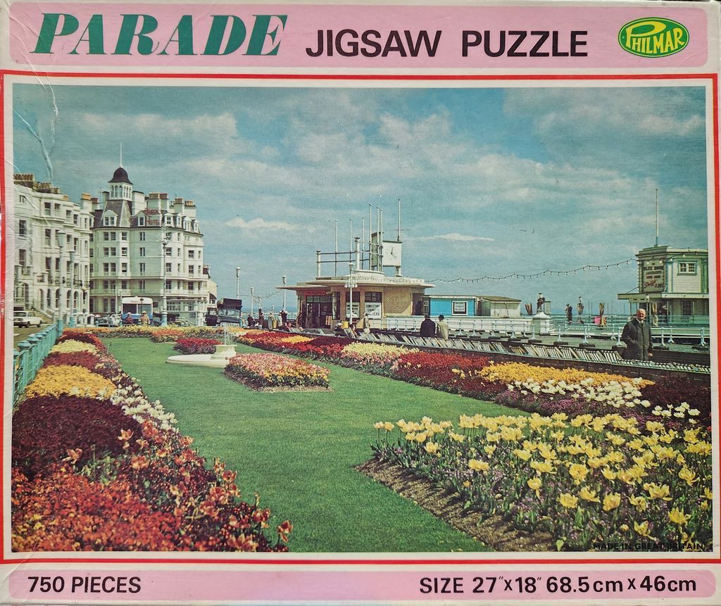 A vintage seaside town square featuring vibrant flower beds, manicured lawns, and grand Victorian-style hotel architecture.