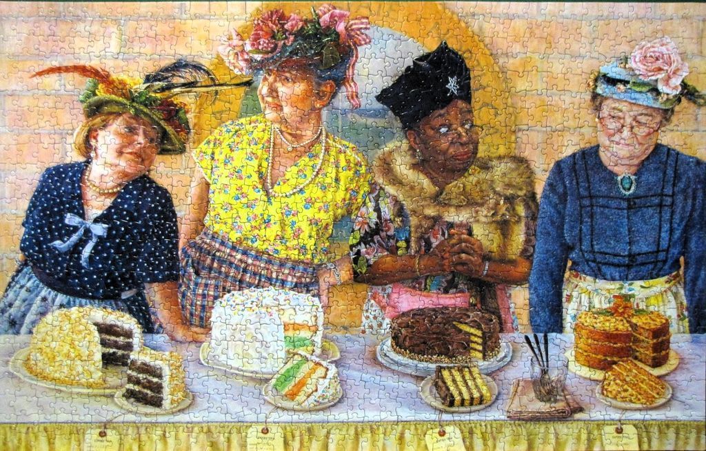 Four women in hats standing behind a table filled with various layer cakes.