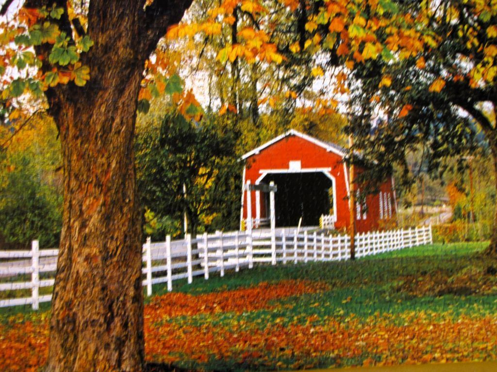 A bright red covered bridge nestled in an autumnal forest with a white picket fence in the foreground.