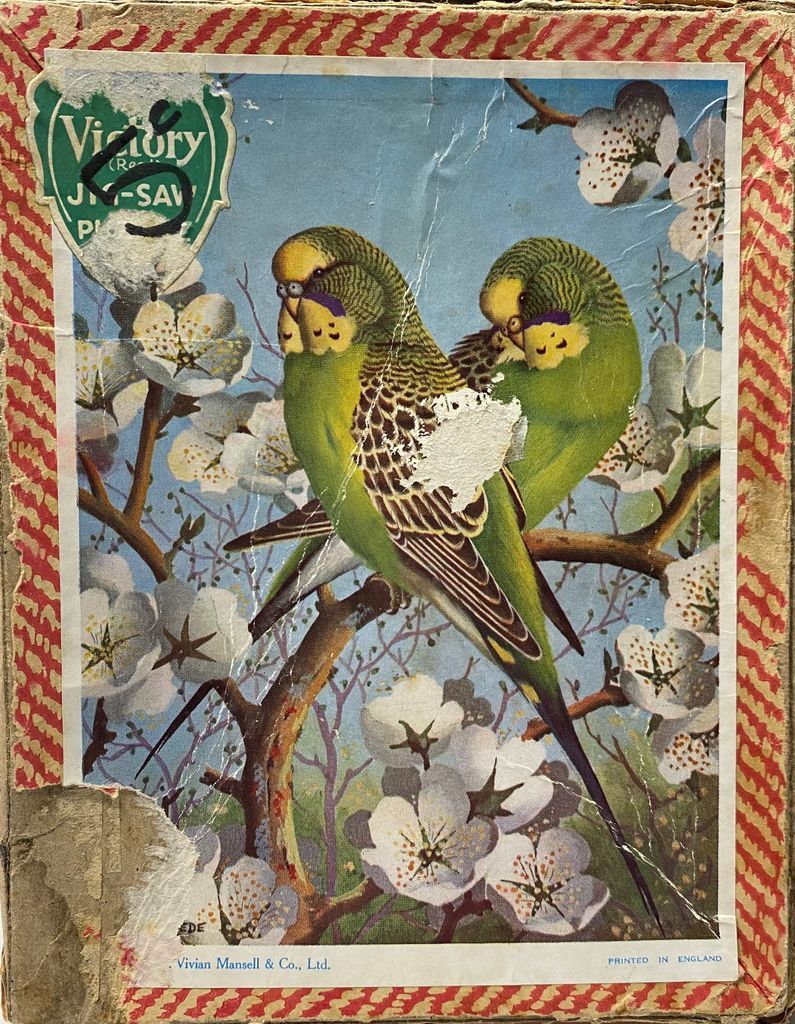 Two green budgerigars perched on a flowering tree branch against a pale blue sky.