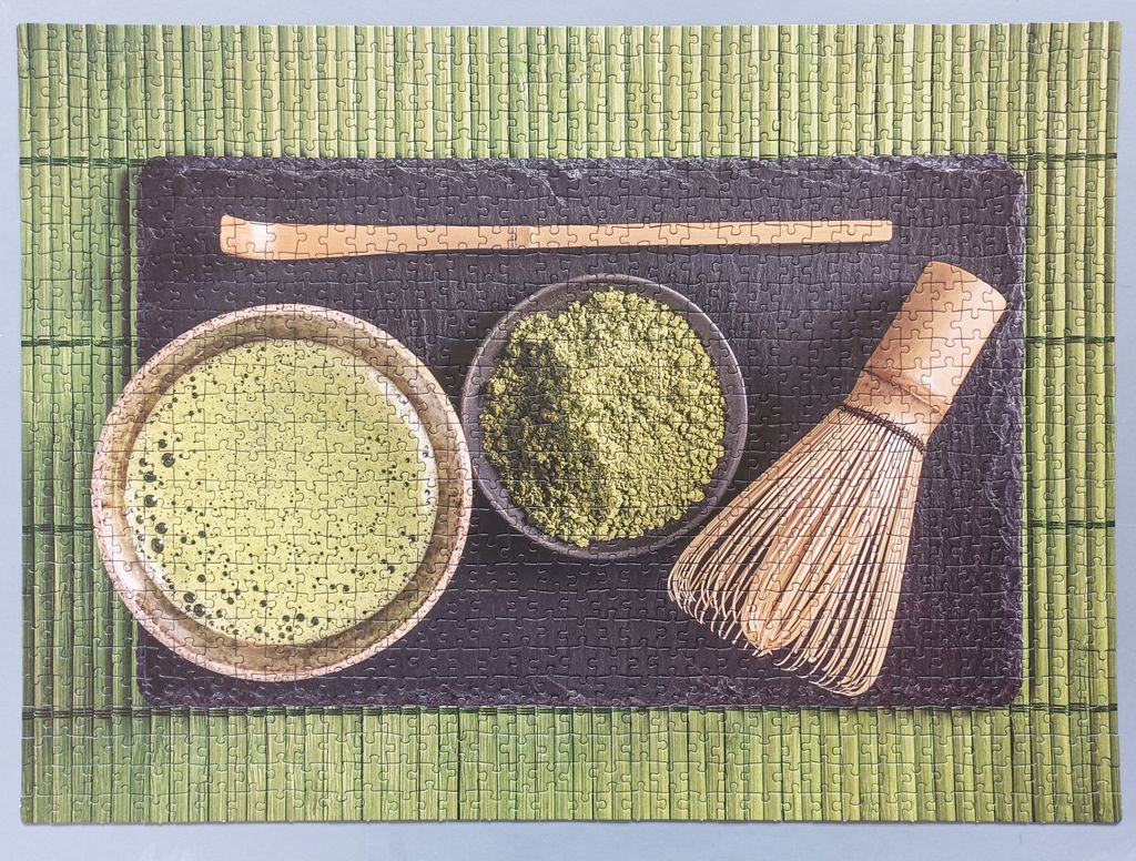 A top-down view of a matcha preparation set with tea, powder, a whisk, and a bamboo scoop on a green mat.