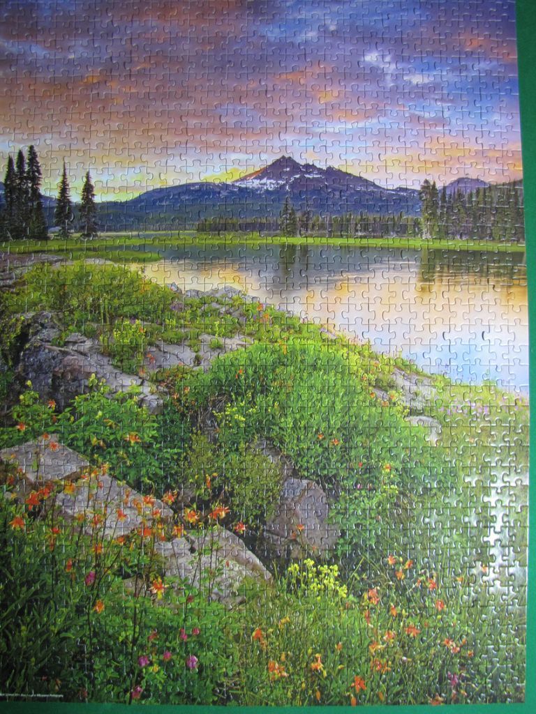 Mountain landscape at sunset reflected in a still lake, with wildflowers and rocky terrain in the foreground.
