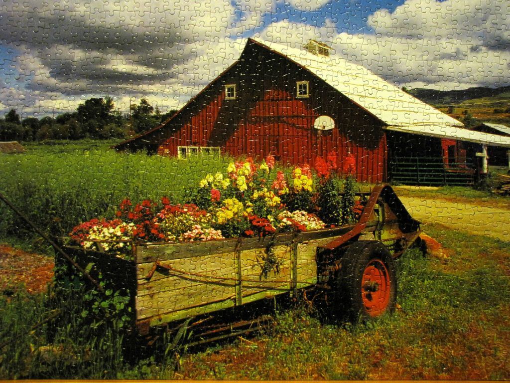 A red barn in a field with a farm wagon overflowing with vibrant flowers in the foreground.