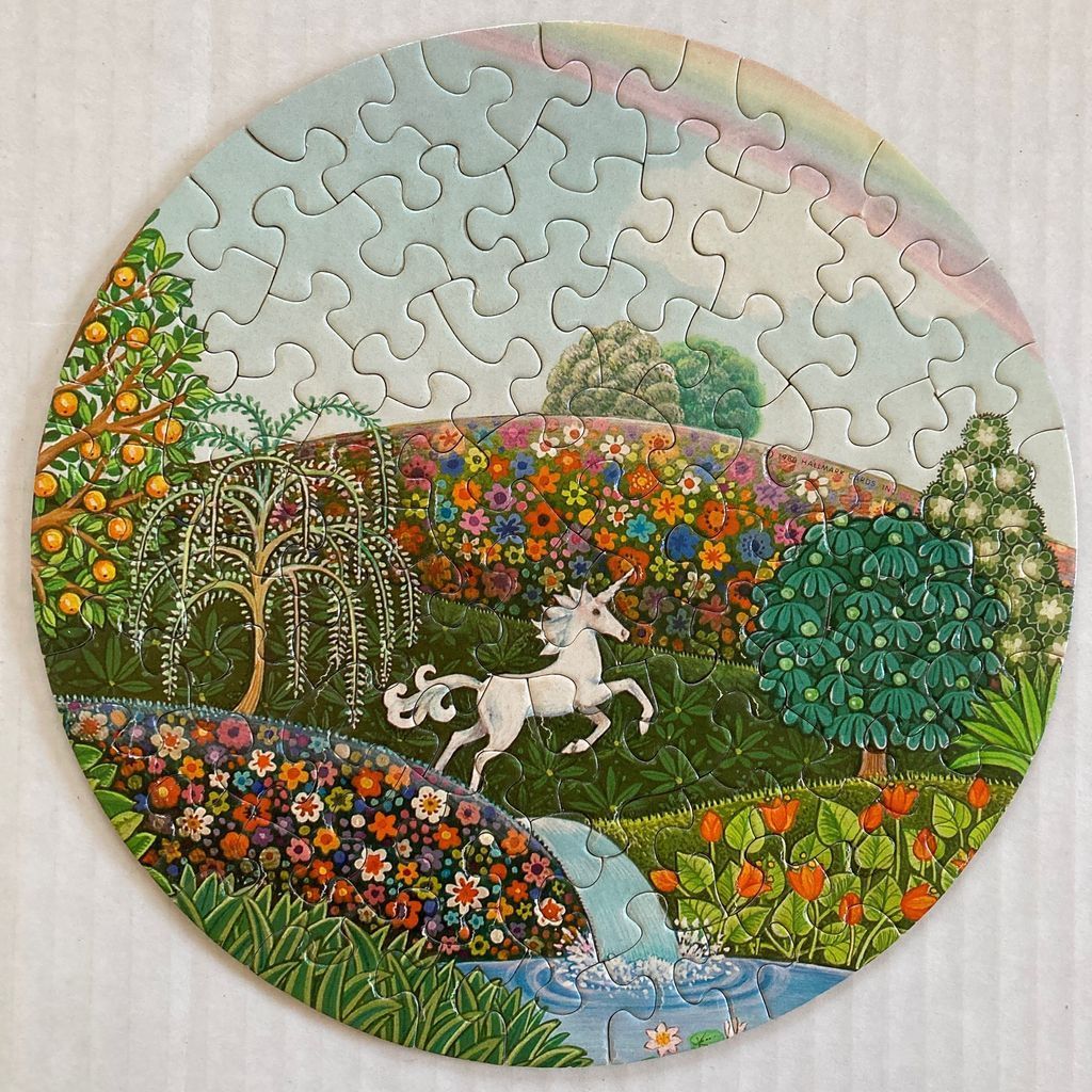 A fantasy illustration of a white unicorn in a lush, floral garden with a stream and a rainbow.