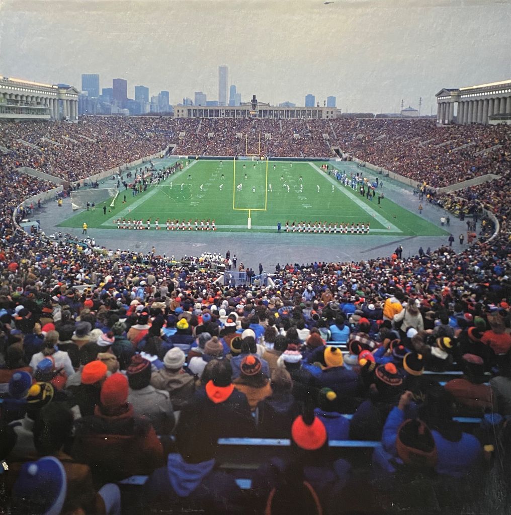 Wide-angle view of a full football stadium, Soldier Field, with a cheering crowd and a city skyline in the background.