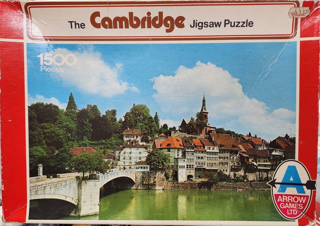 A riverside European village scene with a stone bridge and church steeple under a bright blue sky.
