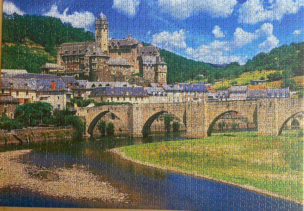 A stone bridge arches over a river in front of a historic European village and castle nestled in a valley.
