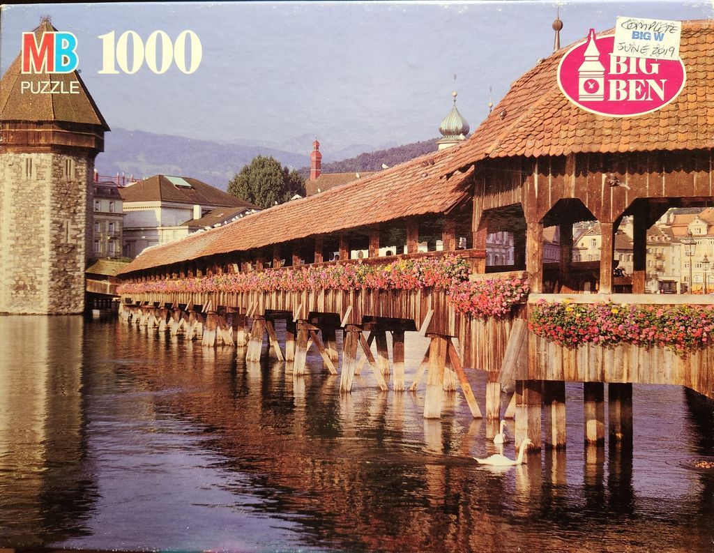 The Chapel Bridge in Lucerne, Switzerland, with flower-lined wooden trusses reflecting in the water at sunset.