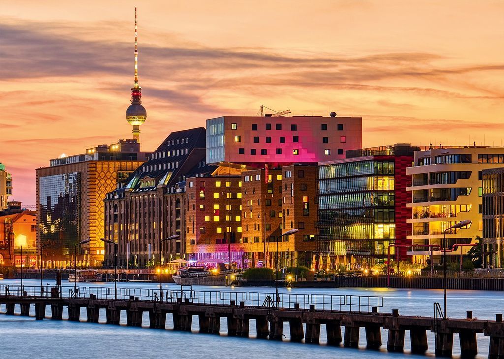 A riverside cityscape in Berlin at sunset featuring the iconic TV Tower and modern architectural buildings.