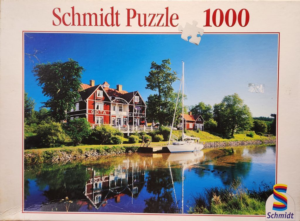 A sunny photograph of the red wood Göta Hotell situated along a calm canal with a sailboat moored at the dock.