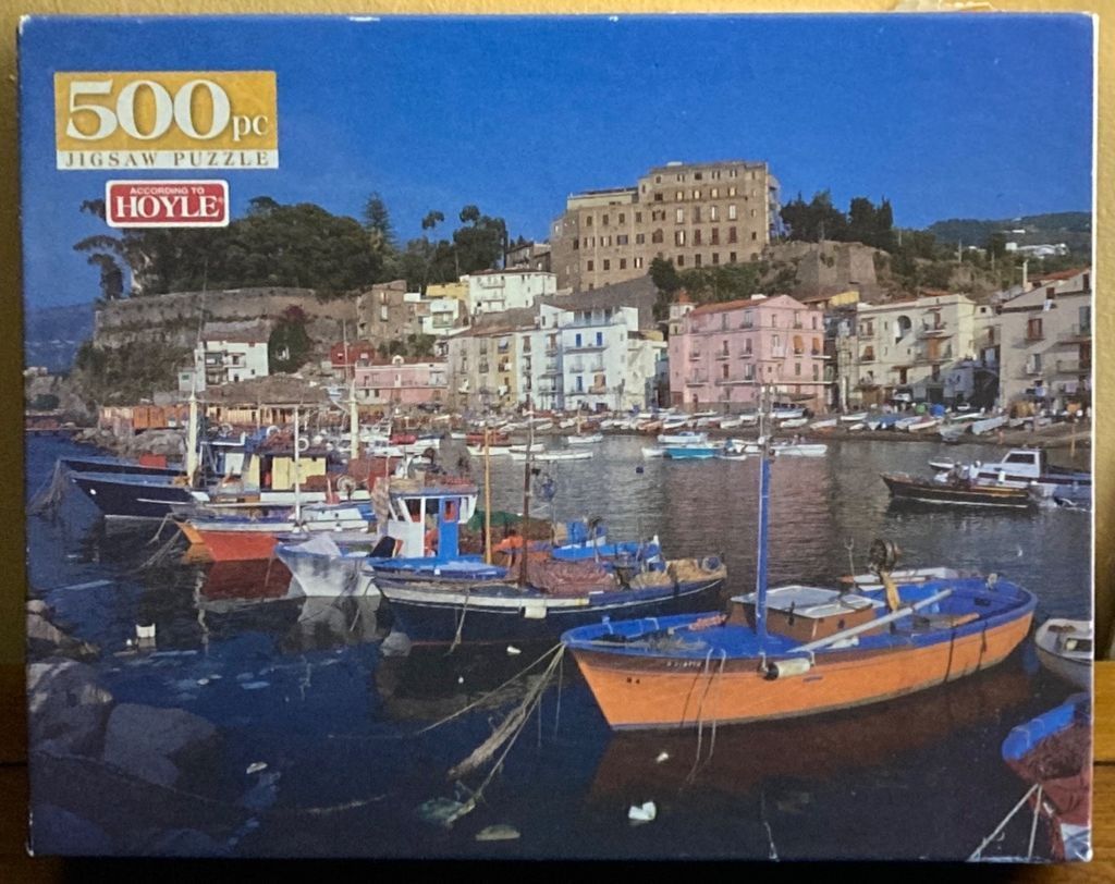 A coastal Mediterranean harbor scene with colorful fishing boats moored in the foreground and town buildings behind.