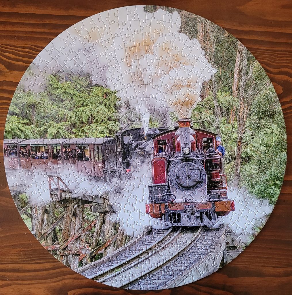 A circular photograph of a vintage steam train traveling along a wooden trestle bridge through a lush green forest.