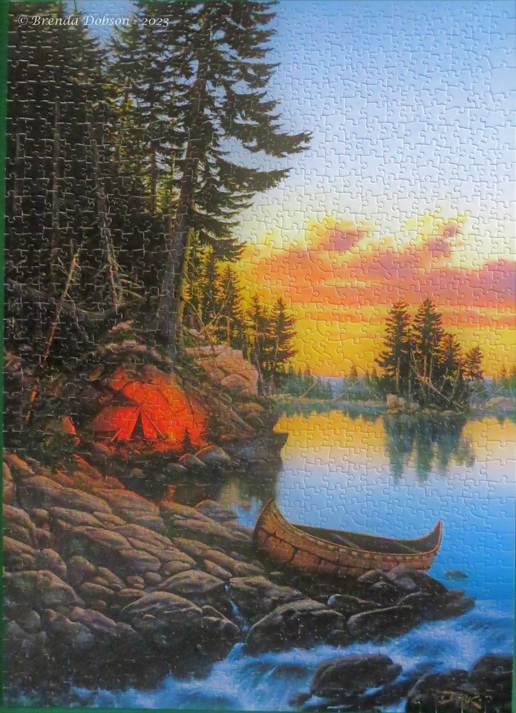 An illuminated tent and a canoe on a rocky shoreline at sunset by a calm lake.