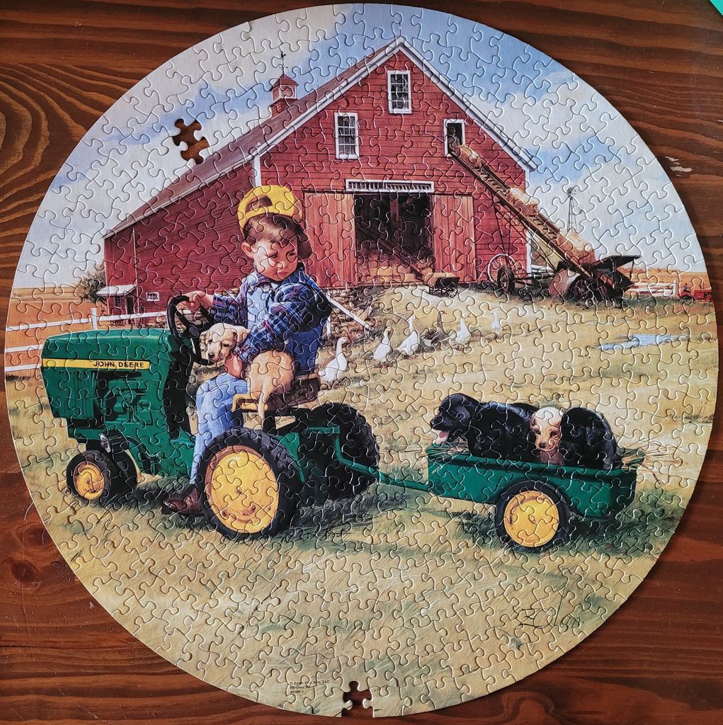 A young child driving a small green tractor pulling a wagon of puppies in front of a red barn.