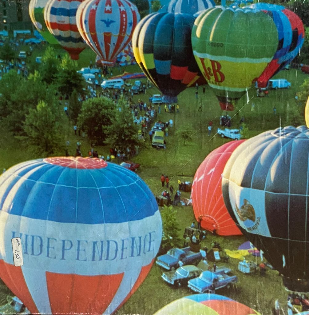 An aerial view of a hot air balloon festival with colorful balloons floating above a grassy park filled with spectators.