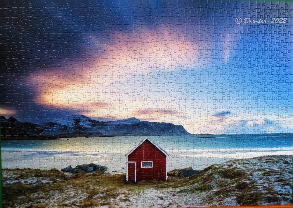 A red cabin stands on a snowy shoreline before a bay, mountains, and a sunset sky.