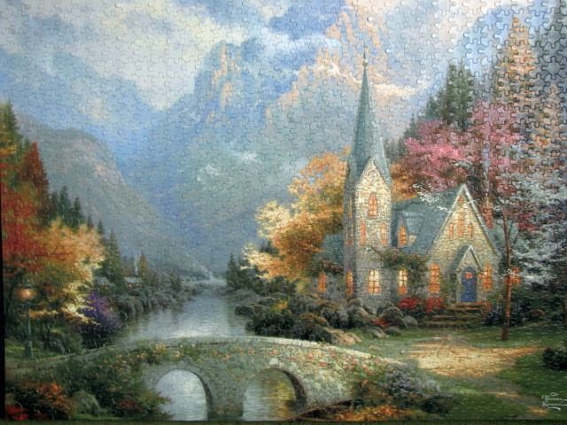 A peaceful stone church by a river and a stone bridge, set against towering mountains in autumn.