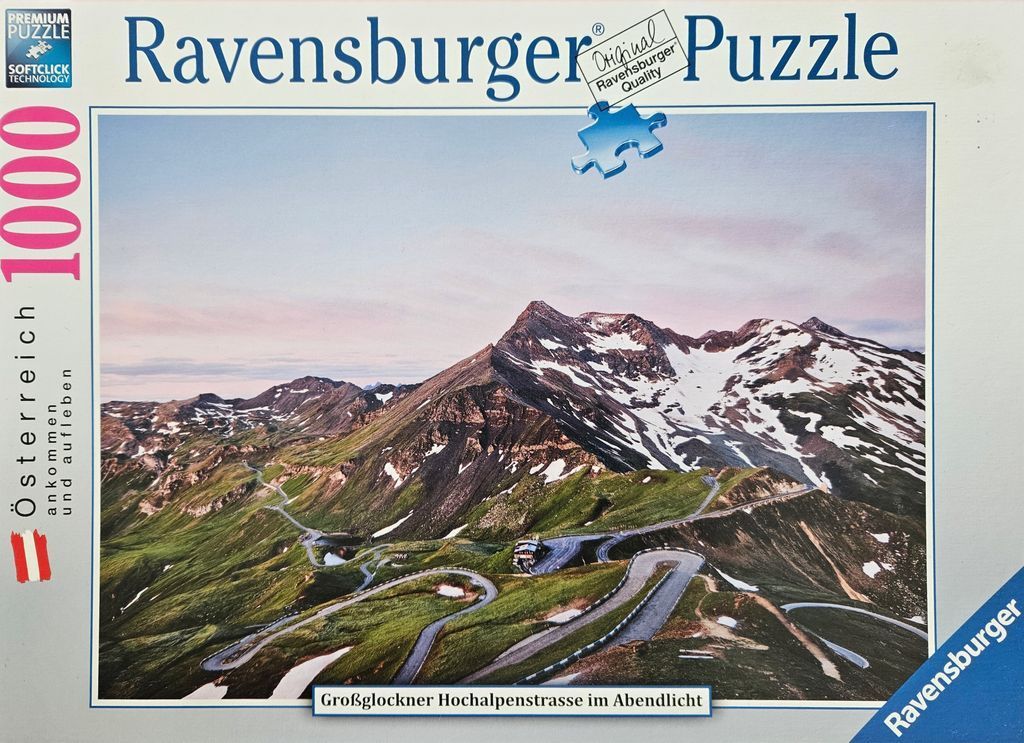 A photograph of the winding Grossglockner High Alpine Road leading up toward snow-capped mountain peaks at twilight.