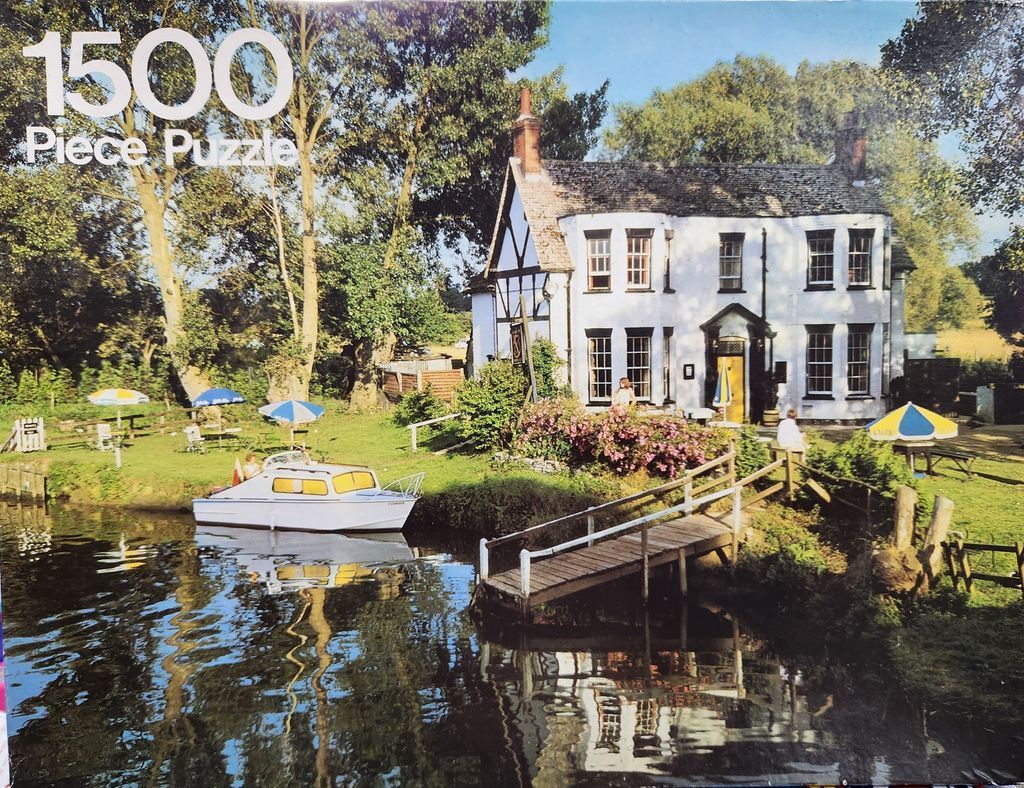 A riverside inn with a white exterior, a small boat docked at a wooden pier, and picnic tables on a grassy lawn.