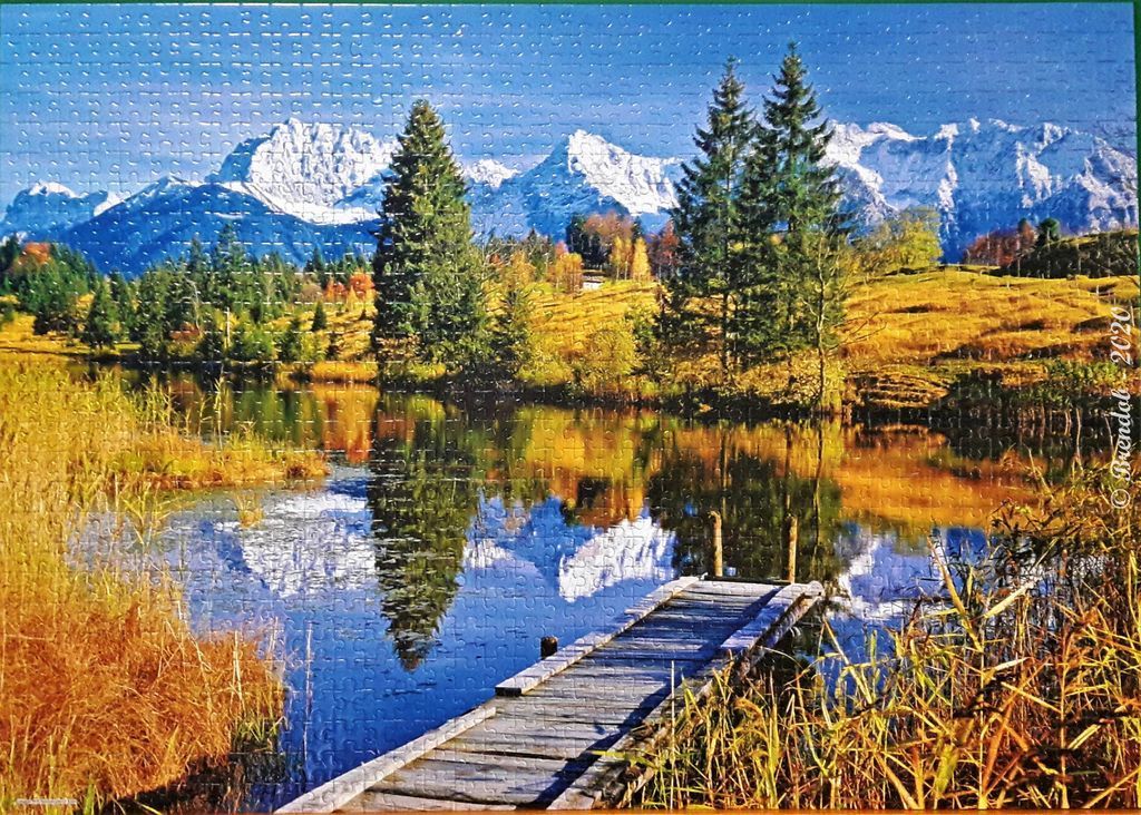 A wooden pier extending into a calm pond reflecting snow-capped mountains and autumn trees.