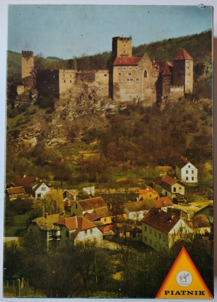 A medieval hilltop castle overlooking a quaint village with red-roofed houses in a valley.