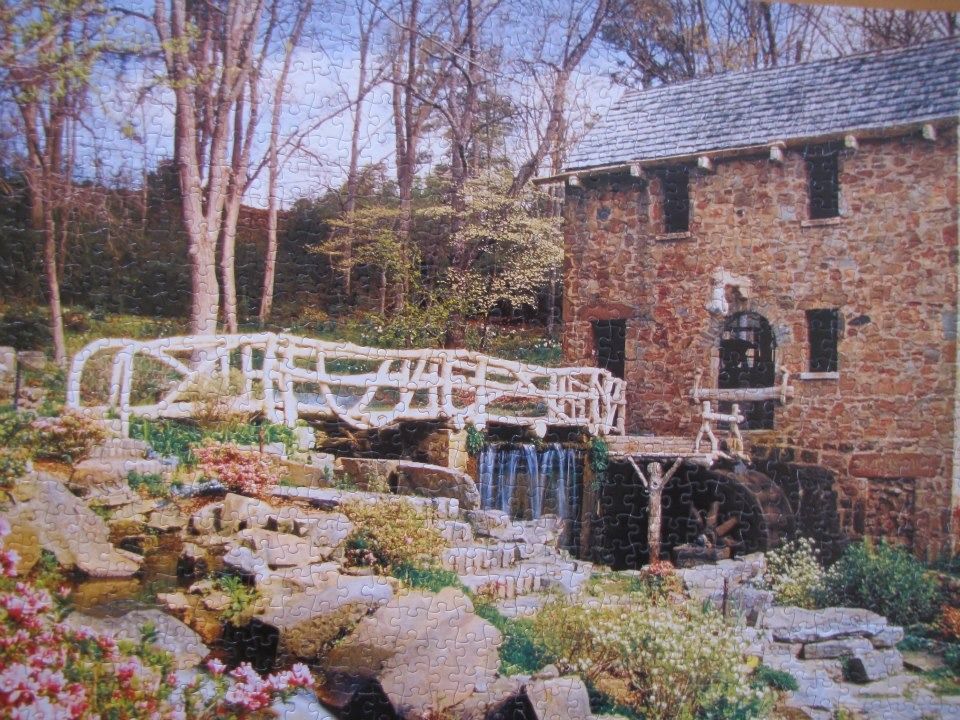 An old stone grist mill with a rustic wooden bridge over a small waterfall and a vibrant garden in the foreground.