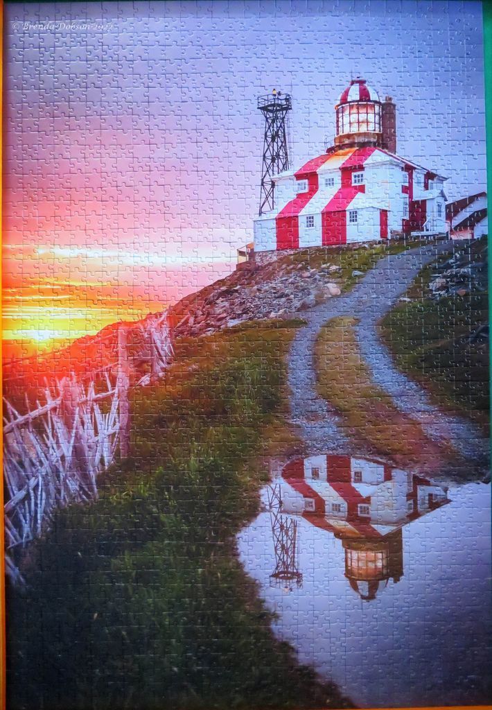 A lighthouse with red and white stripes stands on a hill at sunset, reflected in a puddle on the ground.