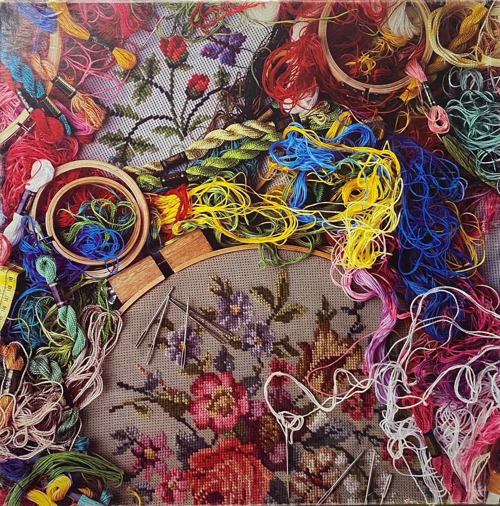 A close-up photograph of vibrant embroidery floss tangled around wooden hoops containing cross-stitch floral patterns.