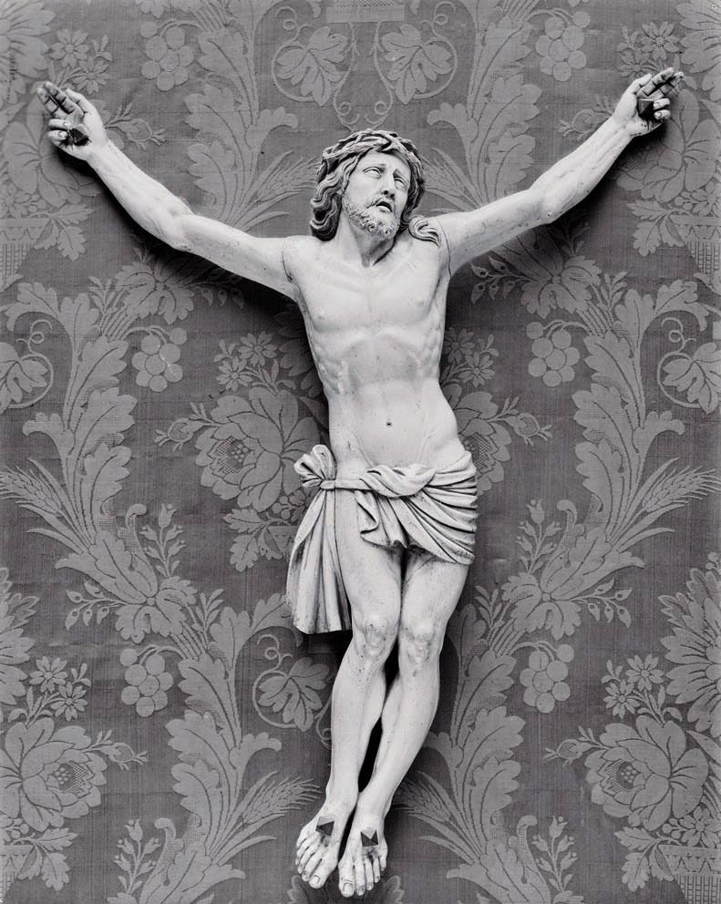 A black and white studio photograph of a carved wooden crucifix against a damask fabric background.