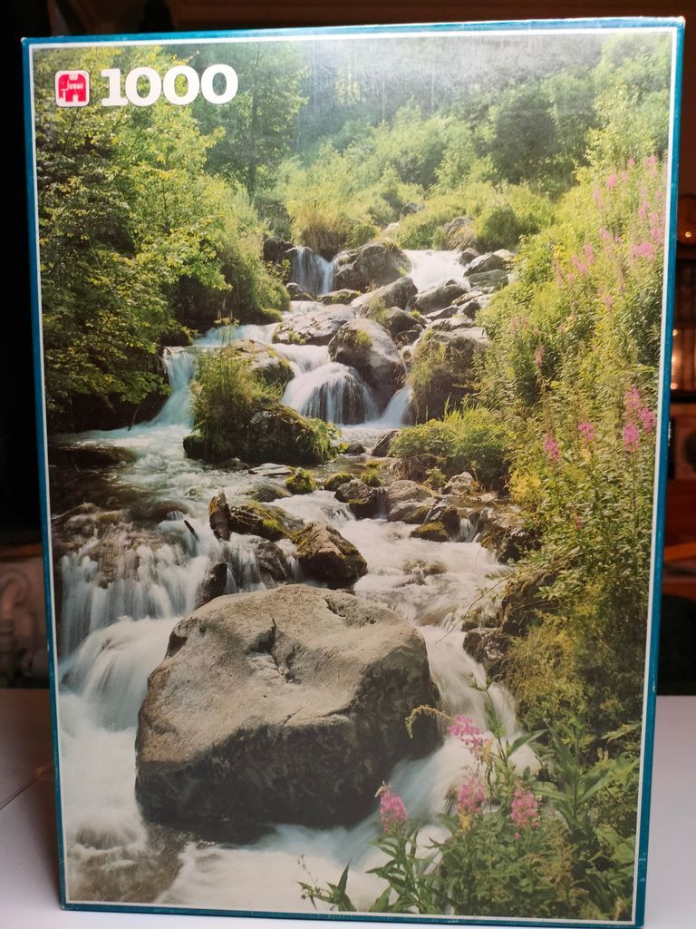 A cascading mountain stream flows over rocks surrounded by lush green foliage and wildflowers.