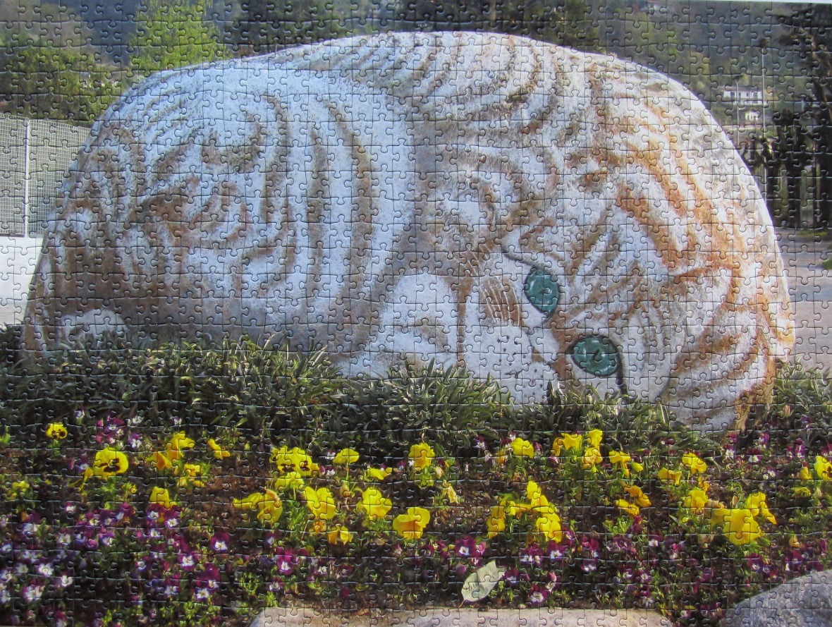 A large, rounded stone painted to resemble a sleeping tiger resting in a flower bed with yellow and purple flowers.