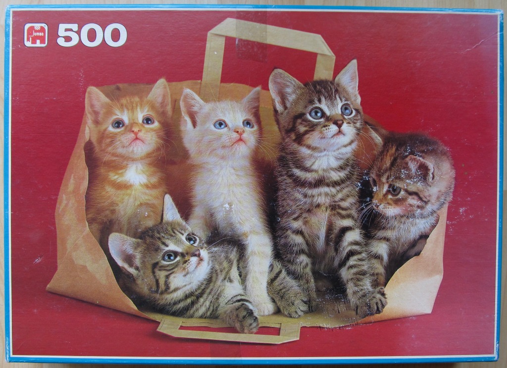 Four kittens sitting and lying inside a brown paper shopping bag against a solid red background.