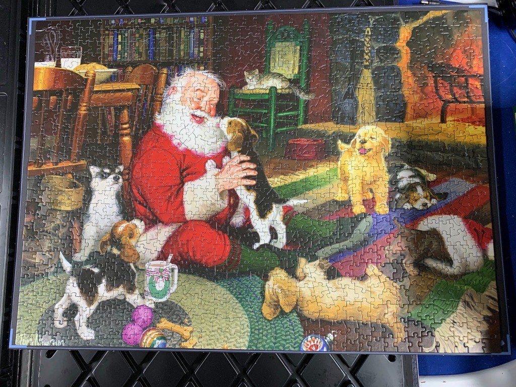Santa Claus sitting on a rug surrounded by playful puppies in a cozy room with a fireplace.