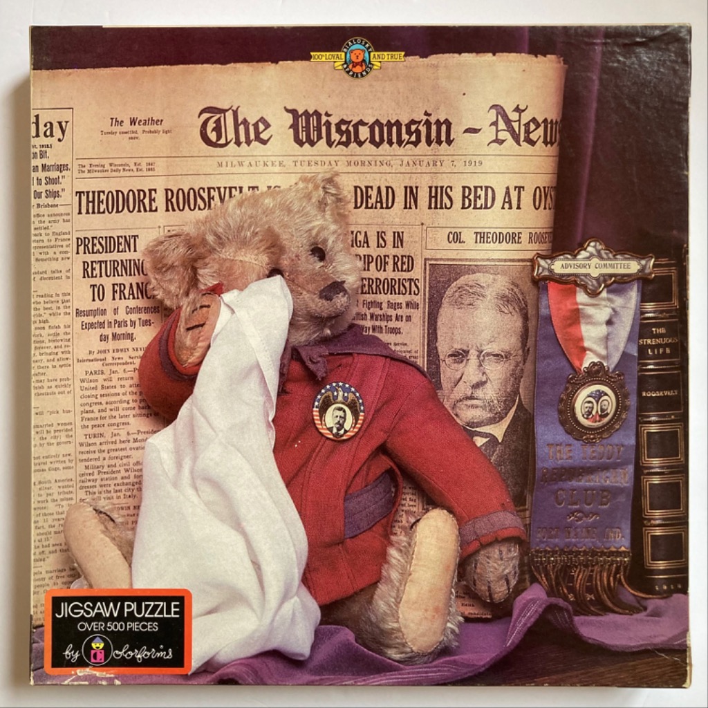 A vintage teddy bear dressed in a red coat holding a white cloth in front of a 1919 newspaper front page.