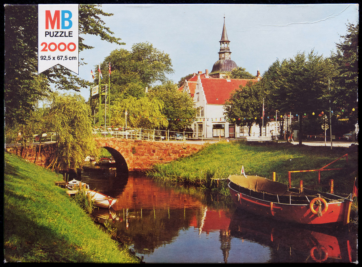 A peaceful canal scene with a stone bridge, red-roofed buildings, a church tower, and a red boat in the foreground.