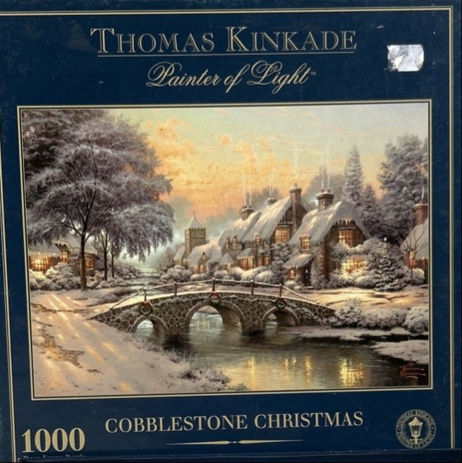 A festive, snow-covered village scene with a stone bridge over a creek at sunset.