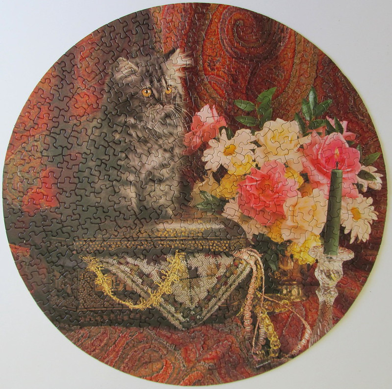 A long-haired grey cat sitting on an ornate box next to a bouquet of flowers and a candle in a round frame.