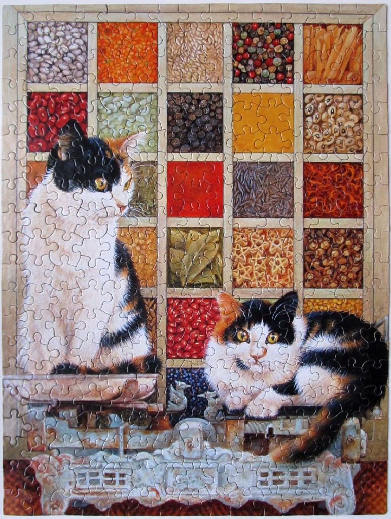 Two calico cats sitting on kitchen scales in front of a spice rack display.