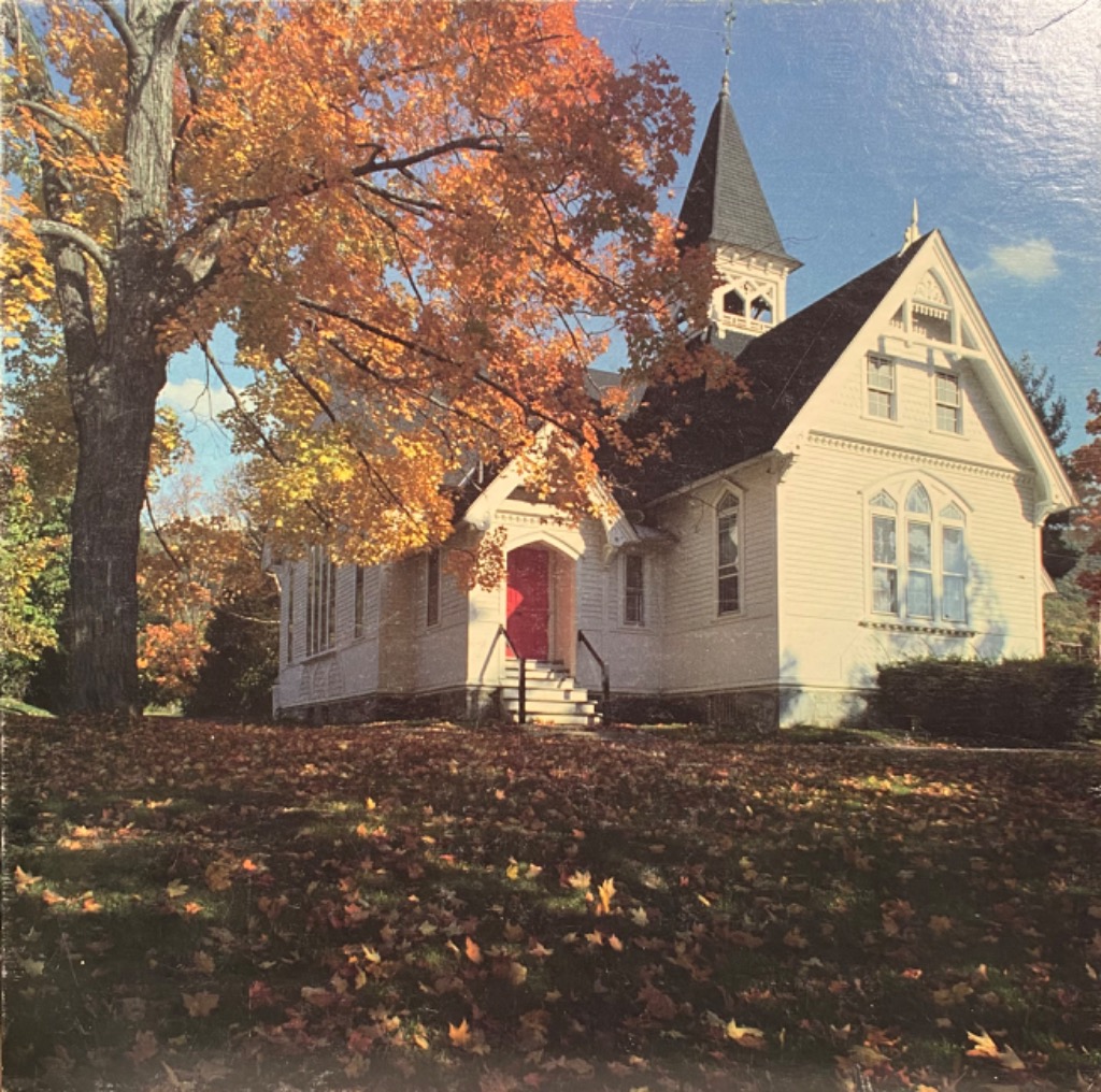 A white country church with a bright red door surrounded by autumn trees and fallen leaves.
